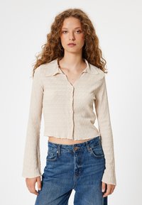 Beige kurzes Hemd mit strukturiertem, gewelltem Design, langen Ärmeln und Knopfverschluss, kombiniert mit blauen High-Waist-Jeans.