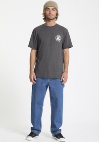 Szary t-shirt z krótkim rękawem z graficznym wzorem na lewej piersi, w połączeniu z luźnymi niebieskimi dżinsami i czarnymi trampkami, na białym tle.