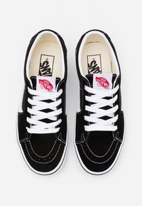 Baskets Vans en daim noir avec des lacets blancs, un logo rouge, et des coutures blanches contrastantes, avec un design bas et une semelle en caoutchouc.