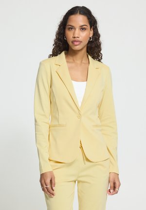 Frau mit lockigem Haar, die einen blassgelben, maßgeschneiderten Blazer und passende Hose über einem weißen Top trägt, vor einem einfarbigen hellen Hintergrund steht.