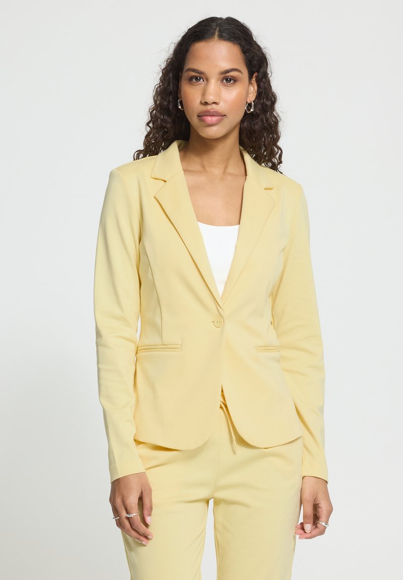 Femme aux cheveux bouclés portant un blazer taillé sur mesure jaune pâle et un pantalon assorti par-dessus un top blanc, debout devant un fond uni clair.