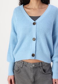 Lichtblauwe gebreide cardigan met een V-hals, lange mouwen en drie bruine knopen. Combinatie met grijze high-waisted jeans. Zachte, gestructureerde stof.