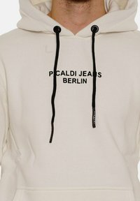 Cream hoodie with black drawstrings and a kangaroo pocket. Features "PICALDI JEANS BERLIN" printed in bold black on the front. Soft texture.