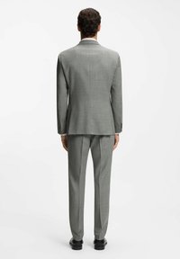 Mężczyzna stojący tyłem, ubrany w jasnoszary garnitur z dopasowanymi spodniami i czarne eleganckie buty na jednolitym białym tle.