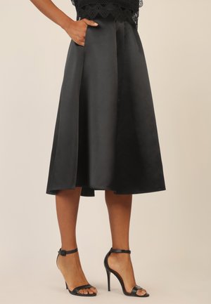 Schwarzer Satin-Midirock mit einem gefältelten Design und Seitentaschen, kombiniert mit schwarzen Riemchen-High-Heels.
