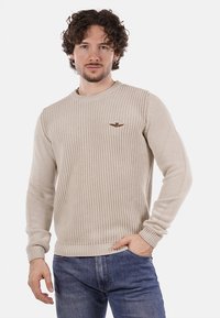 Maglione beige lavorato a maglia con collo rotondo e maniche lunghe. Presenta una trama a costine e un piccolo logo ricamato sul lato sinistro del petto.