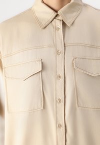 Camicia beige realizzata in tessuto leggero, con chiusura a bottoni sul davanti, colletto a punta e due tasche sul petto con alette angolari.