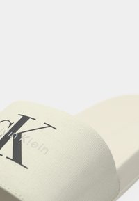 Calvin Klein-skli sandaler i kremfarget stoff, med en fremtredende svart logo, flat design og glatt tekstur med et subtilt mønster.
