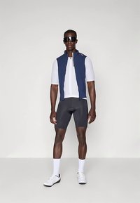 Mannlig syklist iført hvit kortermet trøye, marinblå ermeløs vest, svarte sykkelshorts, hvite sokker, joggesko og store svarte solbriller.