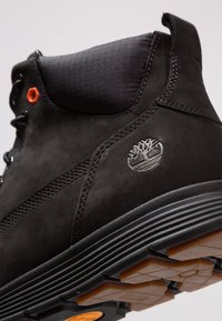 Bottine Timberland noire avec col rembourré, œillet orange, semelle texturée et logo d'arbre en relief sur le côté.