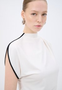 Top bianco con collo alto e maniche corte con rifiniture nere. Tessuto liscio e design aderente. La modella indossa orecchini a cerchio di argento.