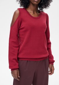 Czerwony dziany sweter z wycięciami na ramionach, prążkowanym dekoltem i mankietami, w luźnym kroju. Noszony z dopasowanymi bordowymi spodniami.