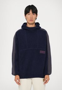 Sweatshirt com capuz em fleece azul marinho, com uma superfície texturizada, riscas laterais e um rótulo roxo. Apresenta um corte relaxado e um bolso canguru.