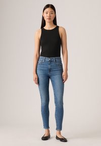 Junge Frau steht, trägt ein schwarzes ärmelloses Top, enge blaue Jeans und schwarze Ballettschuhe vor einem einfarbigen Hintergrund.
