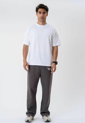 Mand stående, vendt fremad iført en almindelig hvid t-shirt, mørkegrå joggingbukser med tekst, hvide sneakers og et sort armbåndsur.