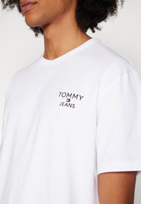 Bílý bavlněný tričko s malou vyšitou logem "TOMMY JEANS" na levé části hrudi, s kulatým výstřihem a krátkými rukávy.