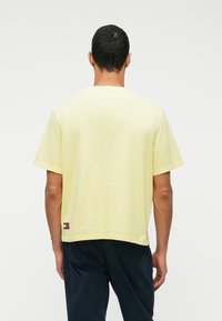 Trumpas rankovės, laisvo kirpimo geltonas marškinėliai, pagaminti iš švelnios medvilnės. Yra apvalus kaklo iškirpimas ir logotipo etikete kairėje pusėje, arti apačios.