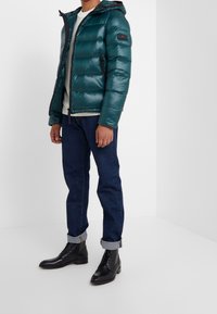 Uomo che indossa un giubbotto imbottito teal, un maglione lavorato a maglia color crema, jeans scuri arrotolati e stivali in pelle nera con lacci, in piedi contro un muro bianco.