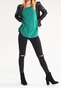 Top verde senza maniche con orlo arrotondato, giacca in finta pelle nera, jeans skinny strappati neri e stivaletti con tacco alto neri.