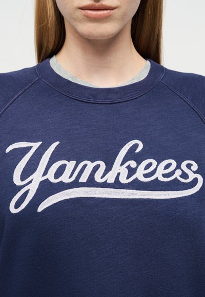 Κοντινό πλάνο ενός ατόμου που φοράει μπλε σκούρο φούτερ με κεντημένο το "Yankees" με άσπρα γράμματα στο στήθος.