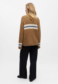 Bruine fuzzy trui met blauwe en witte horizontale strepen, gecombineerd met zwart gestreepte broek. Heeft een ontspannen pasvorm en zijspleten.