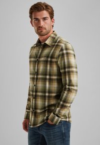 Camicia in flanella a quadri verde oliva, con accenti neri e gialli, colletto classico, maniche lunghe e chiusura con bottoni. Indossata con jeans.