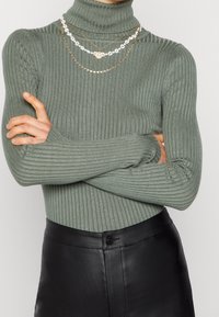 Aansluitende, geribbelde turtleneck trui in gedempt groen. Gelaagd met drie kettingen: twee gouden kettingen en één met een wit bloemenontwerp. Zwarte leren broek.