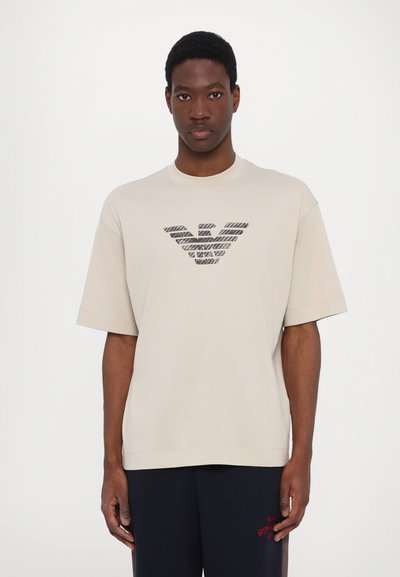 Beige T-shirt met korte mouwen van katoen, met een zwart gestreept logodesign in het midden. Rechtdoor snit met een ontspannen pasvorm.