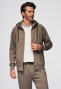 Homme ajustant un sweat à capuche taupe zippé par-dessus un t-shirt blanc, portant un pantalon de survêtement taupe assorti avec un cordon noir sur un fond clair.