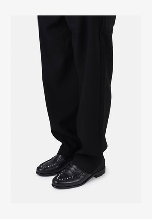 Mustat nahkaiset loafersit, joissa on koristeellisia niittejä ja sileä pinta, yhdistettynä löysiin mustiin housuihin.