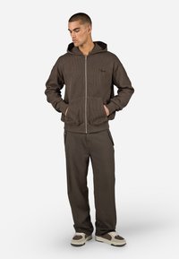 Haut à capuche marron à côtes avec fermeture éclair, doté d'une capuche et de poches avant, associé à un pantalon ample assorti et des baskets crème. Détail de marque simple.