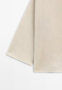 Beige strikket stof med åben vævning, der viser den nedre ærme- og kropssektion af et beklædningsstykke mod en hvid baggrund.