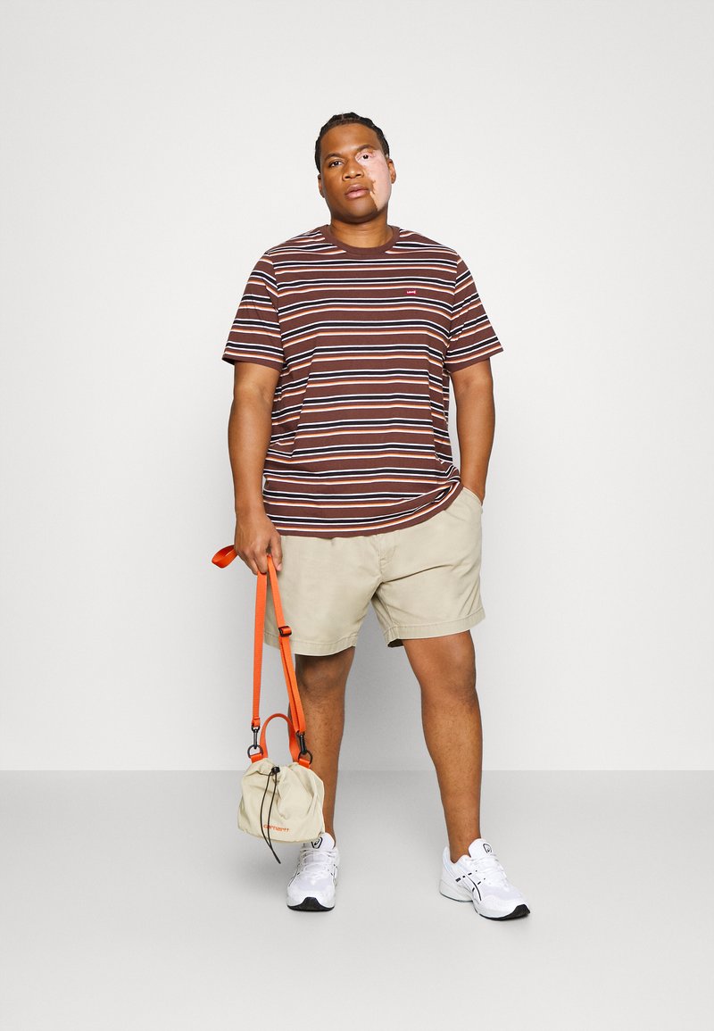 Braun- und cremefarbenspenden gestreiftes kurzärmeliges Hemd, beige Shorts, weiße Turnschuhe, eine beigefarbene Tasche mit einem orangefarbenen Riemen haltend.