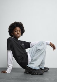 Une jeune personne avec des cheveux afro est assise par terre, portant un haut à manches longues noir et blanc superposé, un jean large bleu clair et des baskets noires.