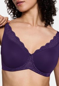 Mulher ajustando a alça de um sutiã roxo com rendas, mostrando o torso superior e cabelo encaracolado, em fundo branco liso.