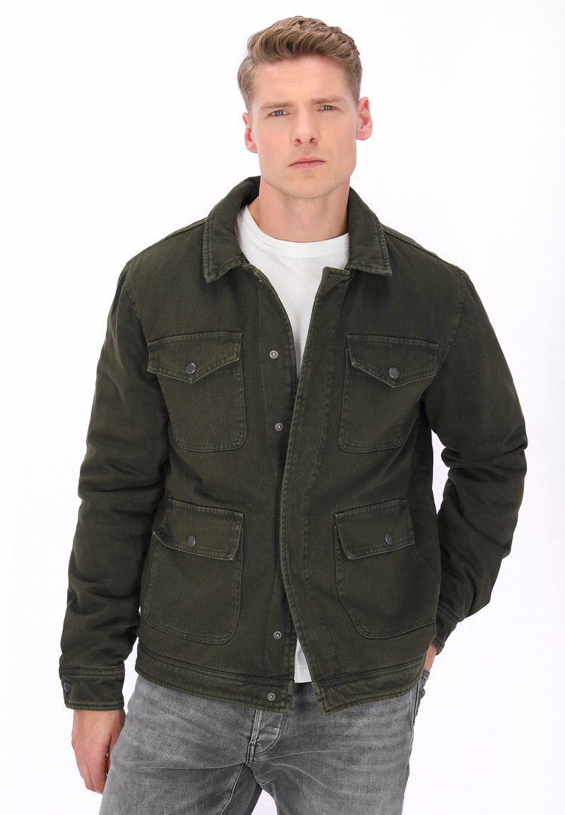 Junger Mann trägt eine dunkelgrüne Jacke mit Knöpfen und vier Klappentaschen, ein weißes T-Shirt und graue Jeans, steht mit einer Hand in der Tasche.