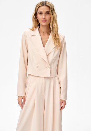 Kobieta o długich blond włosach, ubrana w kremową, krótko skrojoną marynarkę i pasujące, szerokie spodnie z wysokim stanem na białym tle.