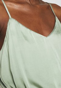 Satin grön cami med V-ringning, tunna axelband och mjukt drapering. Den släta texturen och subtila glansen framhäver designen.