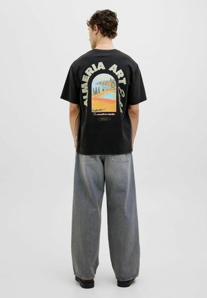 Camiseta de algodón negra con un estampado artístico colorido en la parte posterior. Corte holgado, mangas cortas y combinada con unos vaqueros grises anchos.