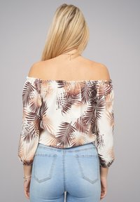 Blusa descubierta en los hombros de tejido ligero con estampado de hojas de palma en marrón, con un escote elastizado y mangas largas y fluidas. Combinada con jeans de un azul claro.