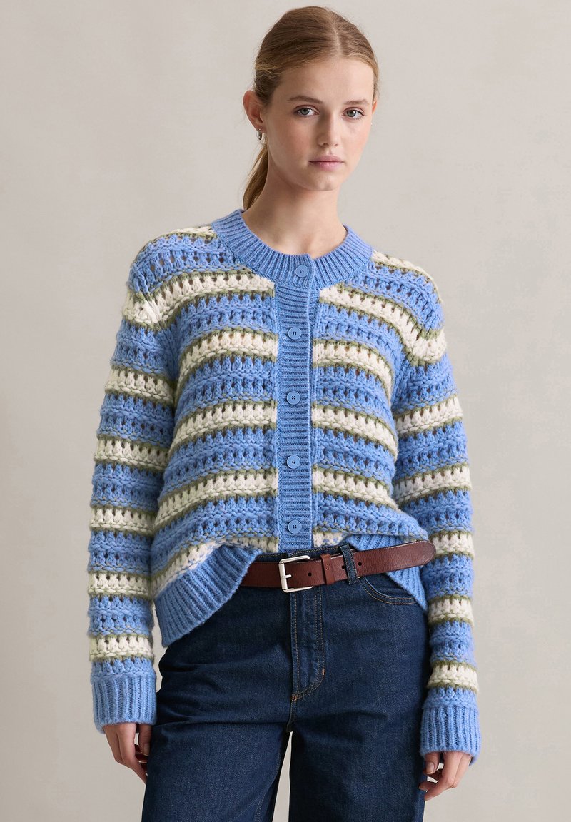 Szydełkowany kardigan w niebieskie, zielone i kremowe paski. Posiada okrągły dekolt, pięć guzików i ściągacze na rękawach. Nosi się go na ciemnoniebieskie dżinsy z brązowym paskiem.