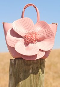 Rosa håndveske med en stor, teksturert blomst som dekorasjon med petalliknende former og intrikate detaljer, plassert mot en trekube.