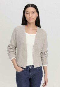 Gebreide cardigan in lichtgrijs, open voorkant, ribbeltextuur, lange mouwen met opgerolde manchetten. Wit t-shirt eronder, gecombineerd met donkere spijkerbroek.