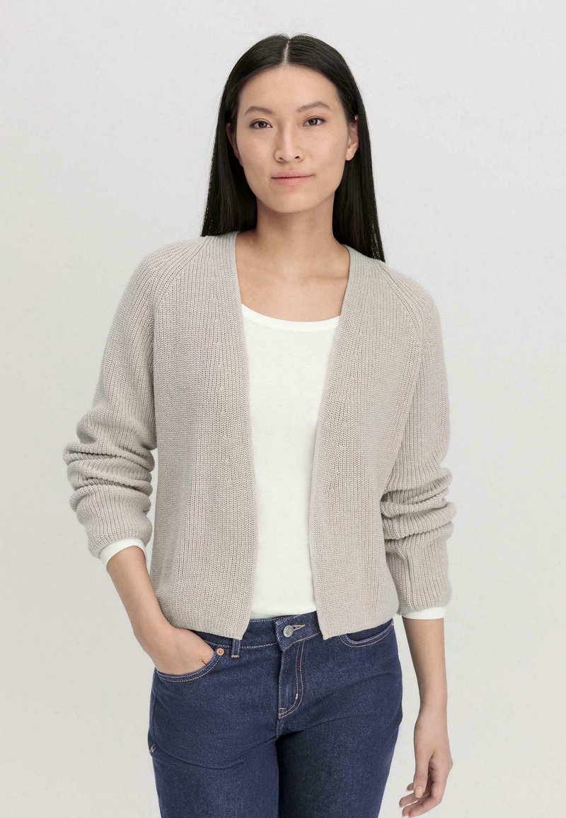 Gebreide cardigan in lichtgrijs, open voorkant, ribbeltextuur, lange mouwen met opgerolde manchetten. Wit t-shirt eronder, gecombineerd met donkere spijkerbroek.