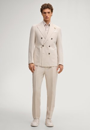 Mann steht mit cremefarbenem zweireihigem Blazer, passenden Hosen, hellem Hemd und cremefarbenen Sneakern vor einfarbig hellem Hintergrund.