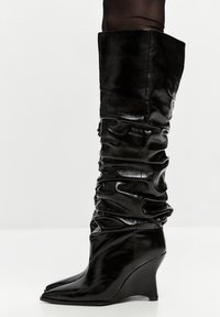Stivali neri al ginocchio realizzati in pelle lucida e testurizzata. Caratterizzati da un design slouchato e un tacco wedge con punta affusolata.