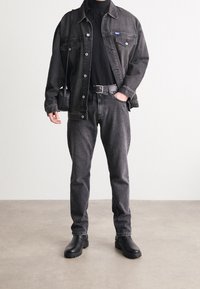 Chaqueta de mezclilla negra sobre un suéter de cuello alto negro, combinada con jeans grises y botas de cuero negras. Un bolso pequeño negro cuelga del hombro.