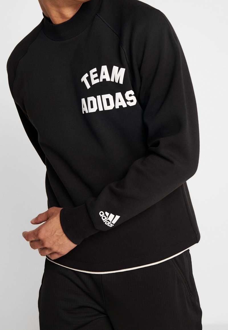 Personne portant un sweat-shirt noir avec le texte blanc "TEAM ADIDAS" sur la poitrine et le logo Adidas sur le poignet gauche.