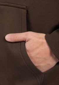 Pravá ruka částečně zasunutá v kapse hnědé dlouhé košile nebo bundy, palec odpočívající venku z kapsy.