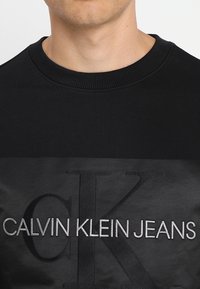 Svart sweatshirt med ribbad krage och strukturerad tyg. Har en framträdande logotyp i en ljusare nyans med texten "CALVIN KLEIN JEANS."